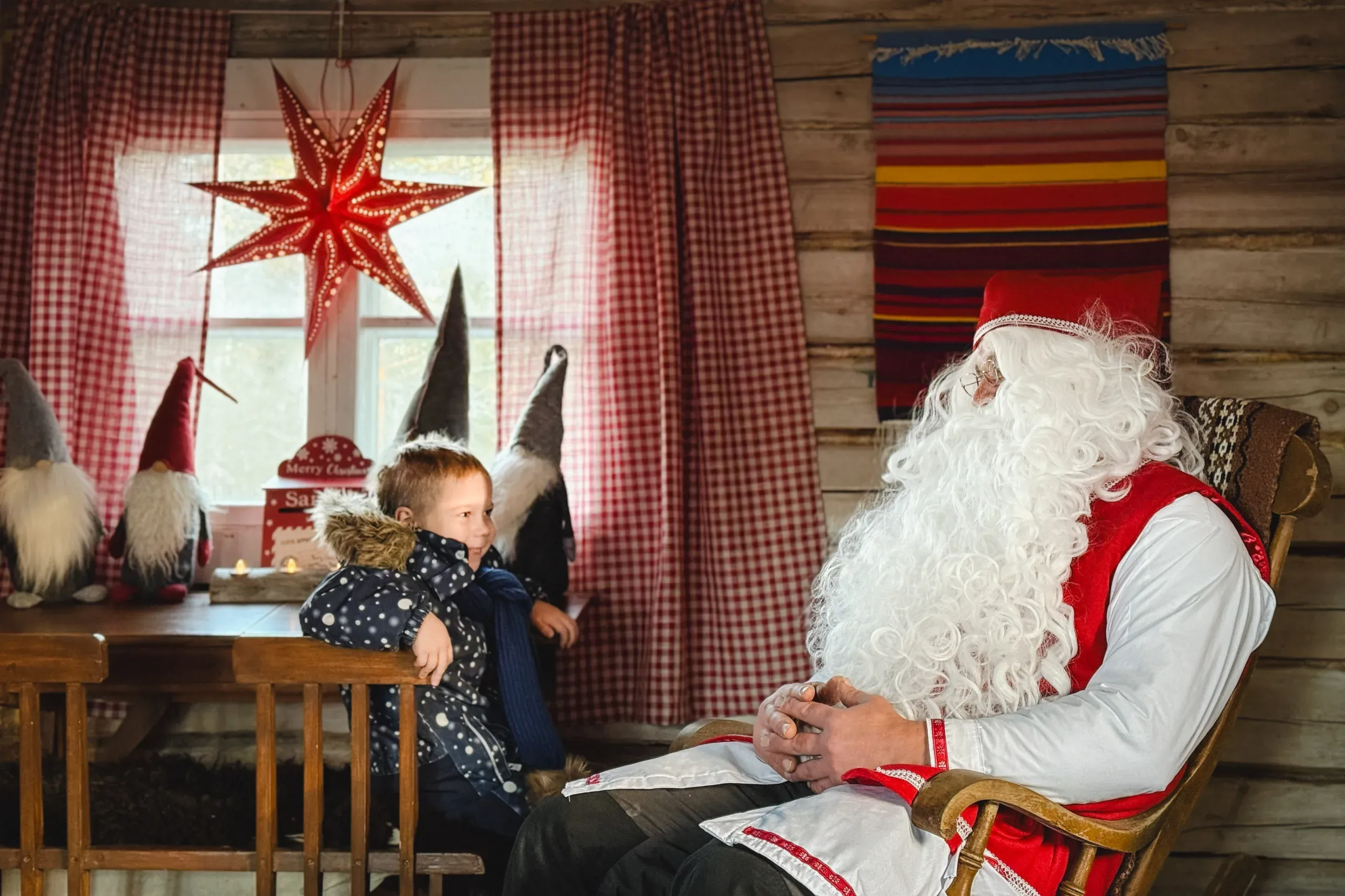 A small boy in a spotted winter coat sitting at a wooden table beside Santa Claus in a red waistcoat, in a log cabin with a red star lamp in the window and felt gnomes on the sill