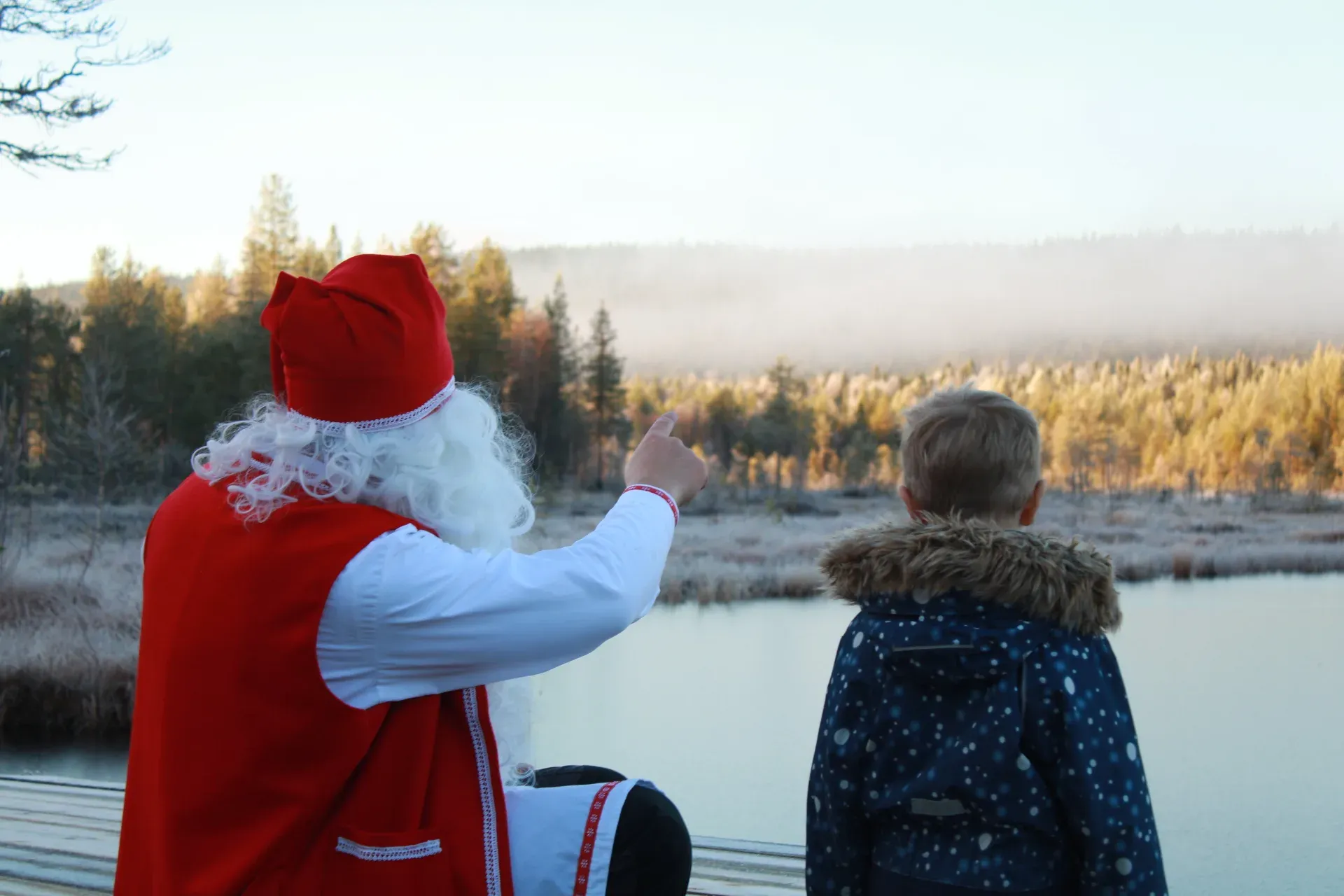 Babbo Natale con il mantello rosso accovacciato accanto a un bambino su un pontile, che indica oltre l'acqua immobile un bosco autunnale brinato