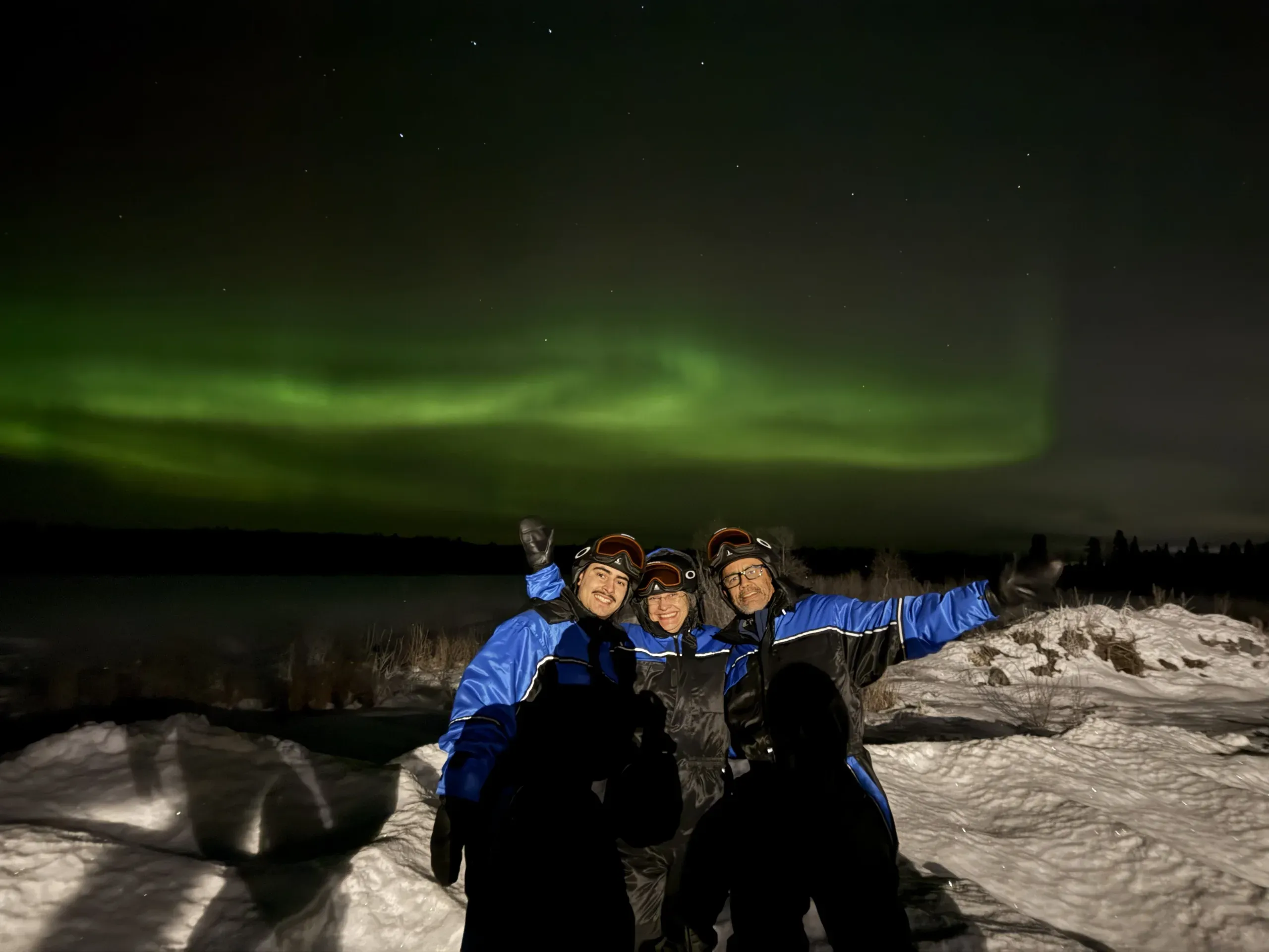 A small group in blue winter overalls and helmets standing close together on snow at night, arms raised, under a band of green aurora over a dark shoreline