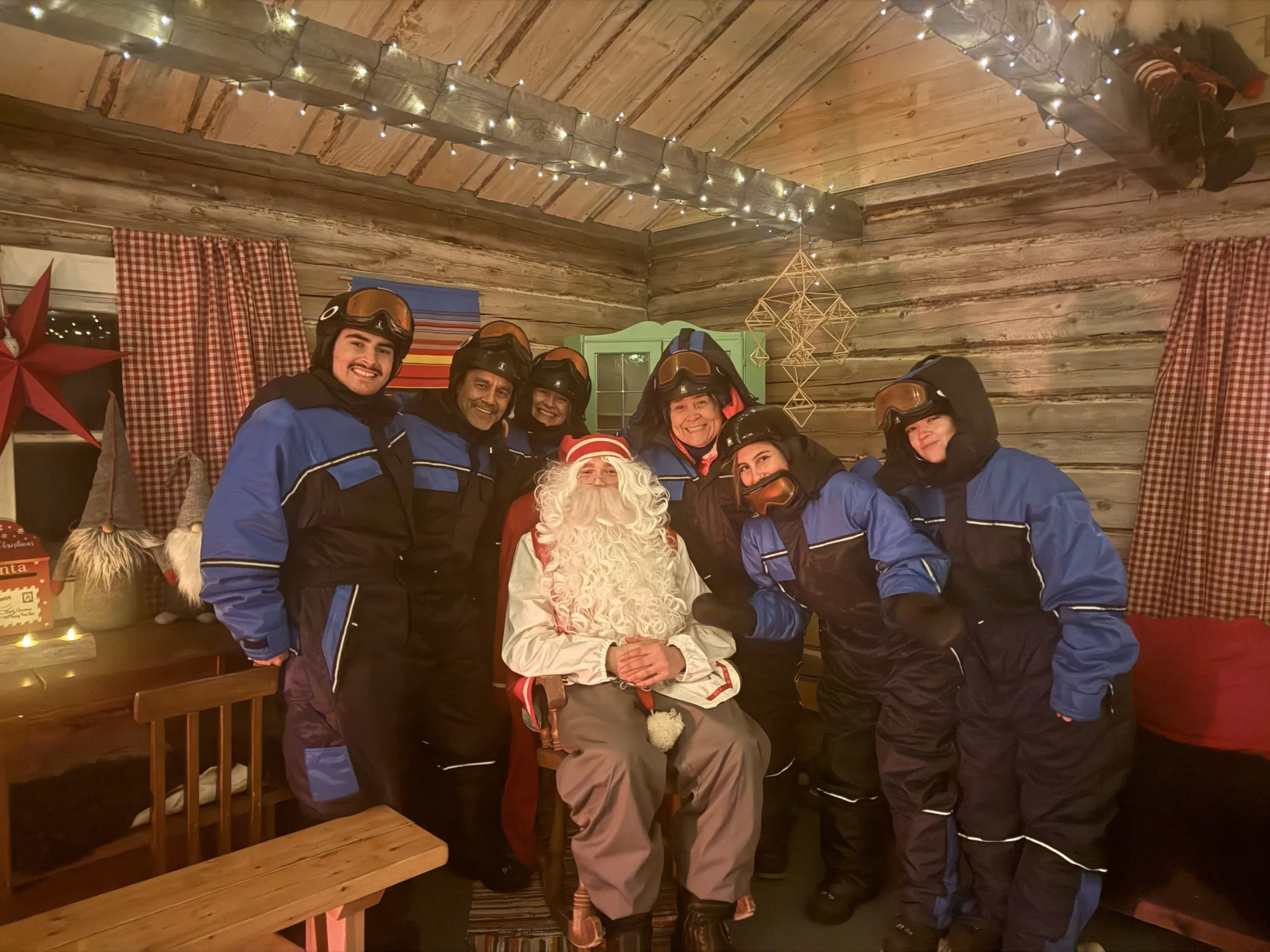 Six visitors in blue thermal suits and helmets crowded around Santa Claus, who sits in a rocking chair in a log cabin hung with fairy lights