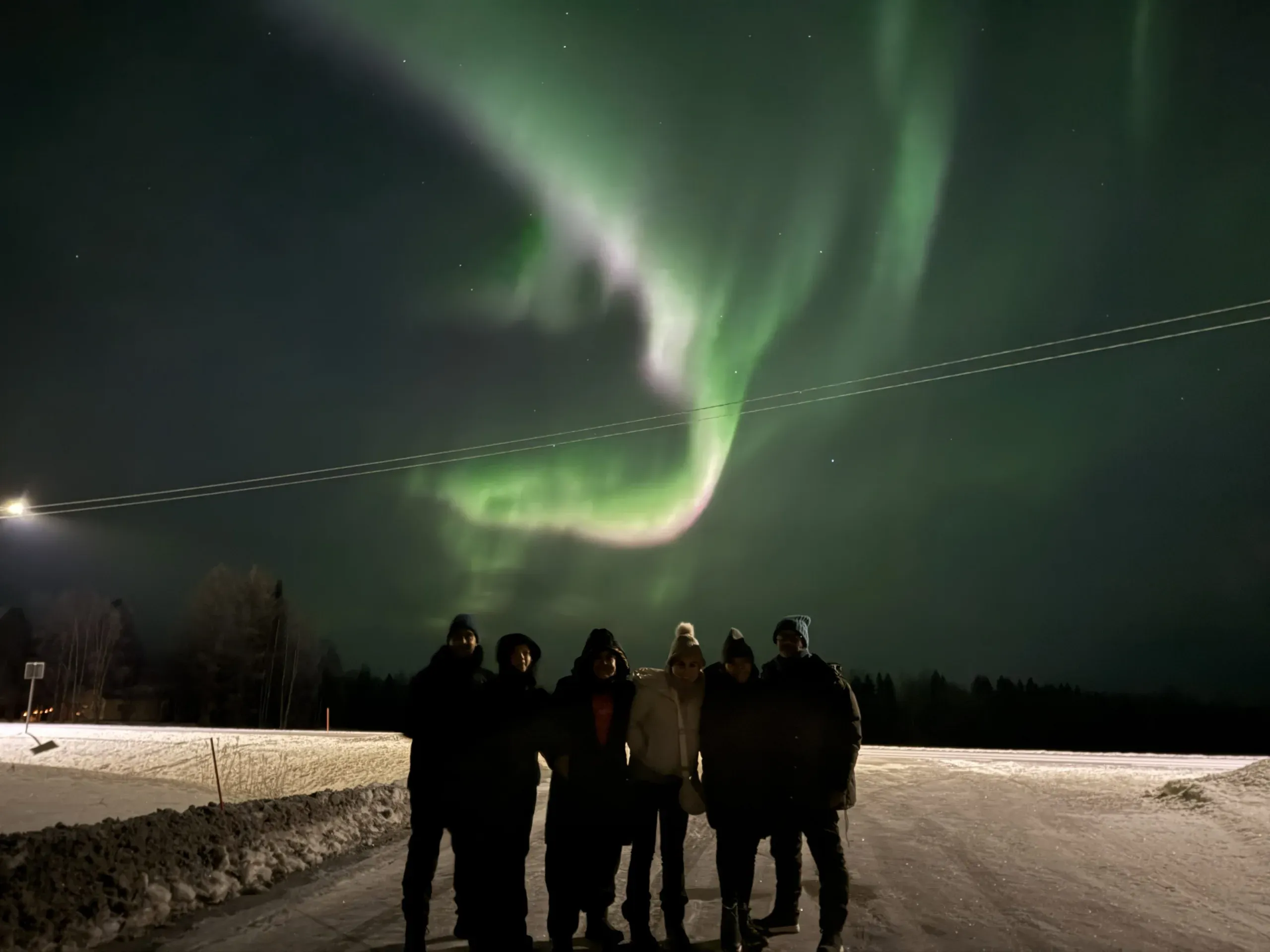 Six people standing as dark silhouettes on a snowy roadside at night under a bright green aurora curling across the sky, a street lamp glowing at the edge of the frame