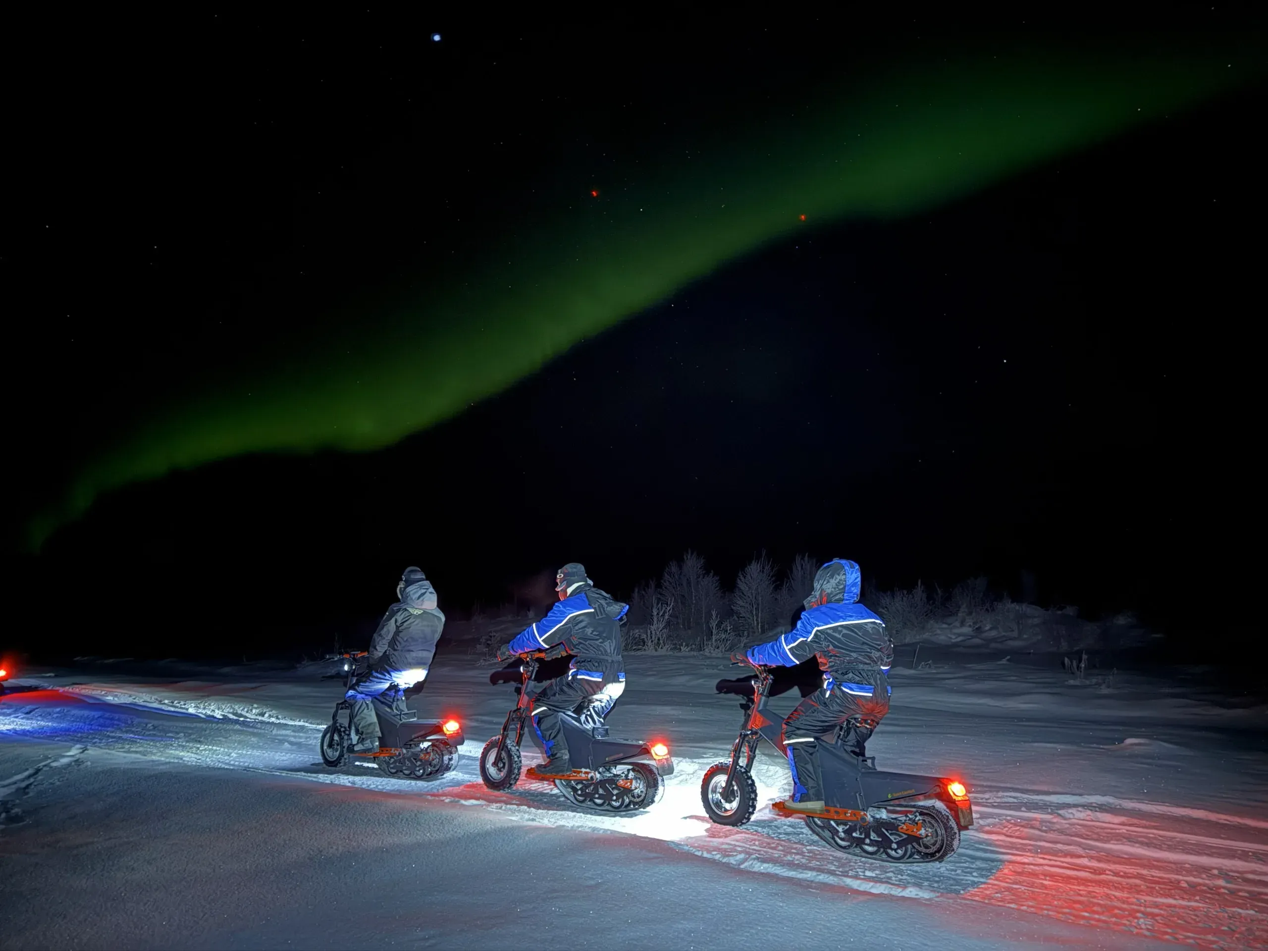 Drei Fahrende auf Elektroschlitten mit Raupen und roten Rücklichtern auf einer beleuchteten Schneespur bei Nacht, darüber ein grünes Nordlichtband am schwarzen Himmel
