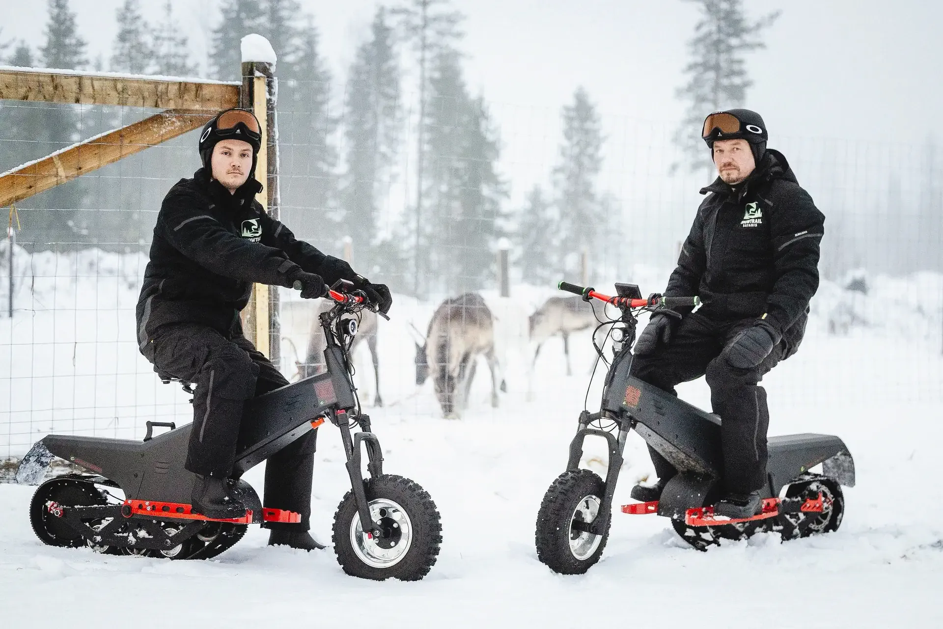 Two guides in black winter gear sitting on tracked electric sleds in front of a wire fence, with reindeer grazing in the snow on the far side and frosted spruce fading into mist