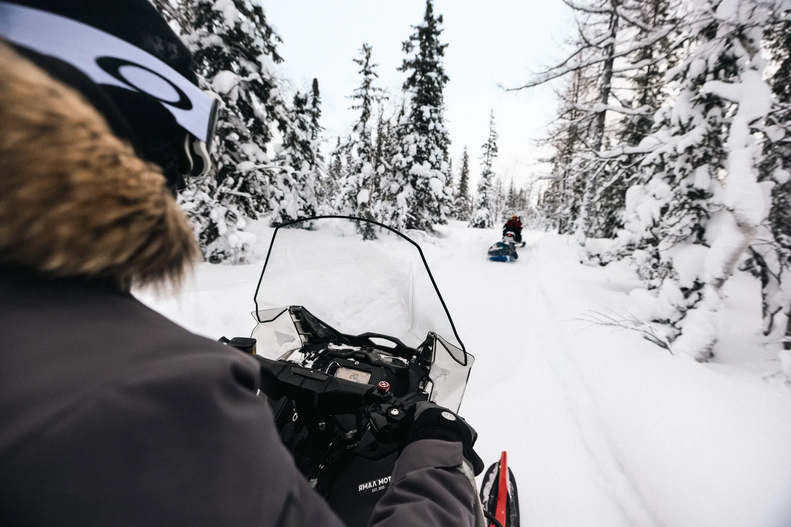 The view over a snowmobile windscreen and handlebars along a deep, narrow trail, a second machine towing a blue sledge some way ahead between heavily snow-laden spruces