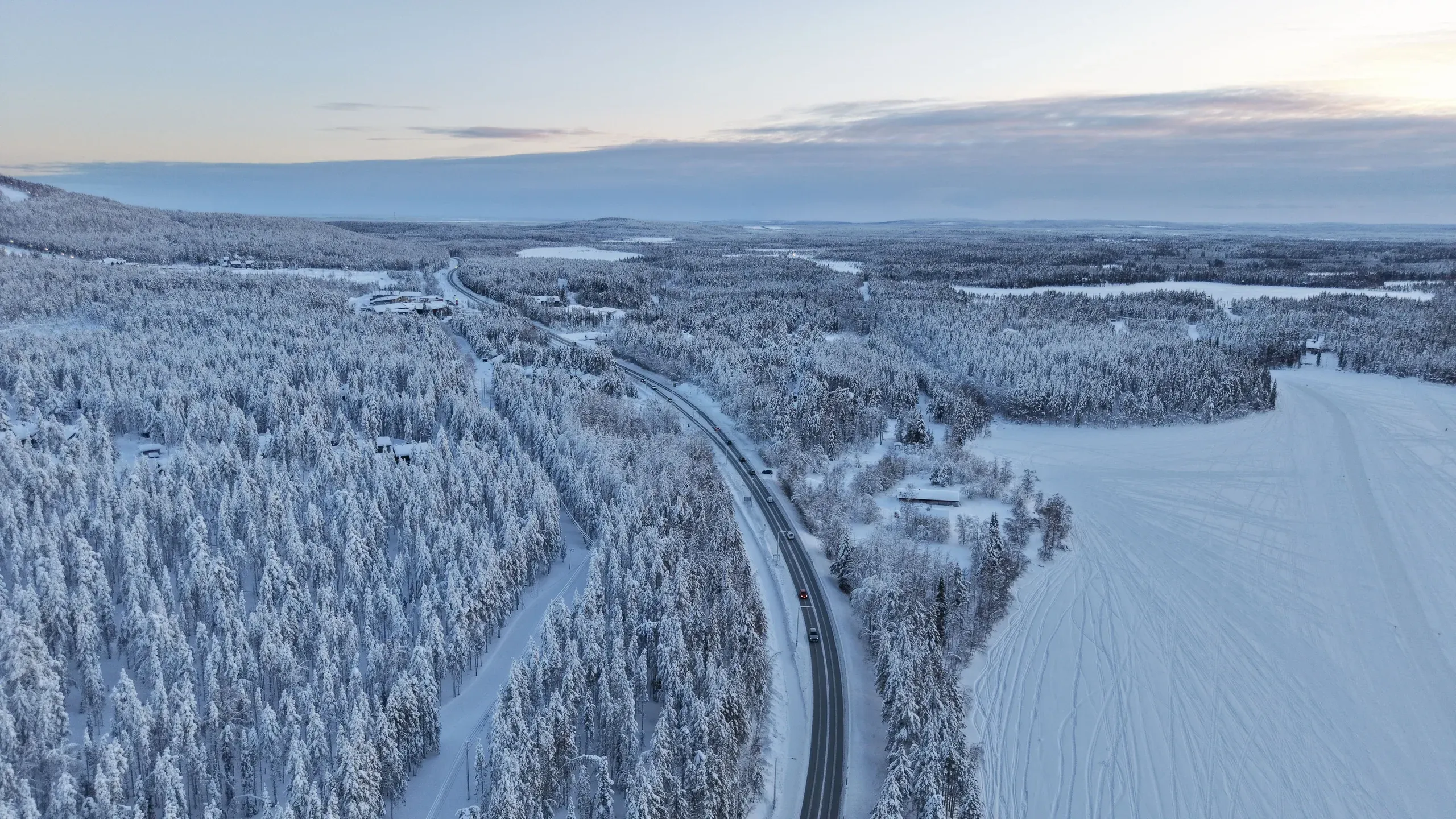 Vista aérea de una carretera despejada que atraviesa un bosque de abetos muy escarchados al anochecer, con edificios dispersos a lo largo y un campo nevado pisado a un lado